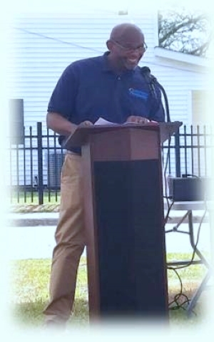 A man in a dark shirt and tan pants speaking at a podium.