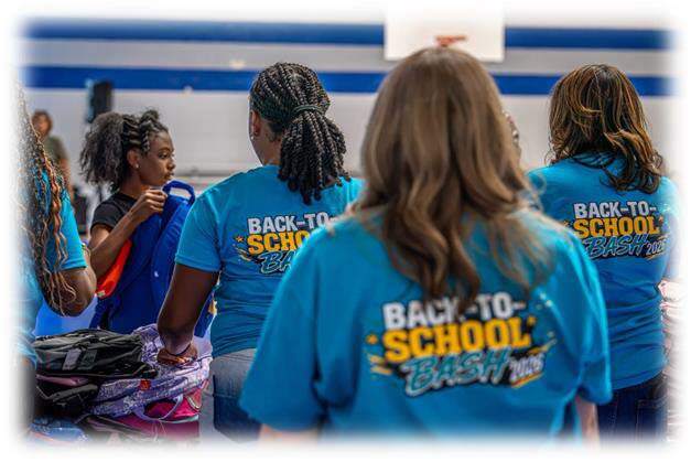 The backs of three Back to School Bash workers passing out a backpack to a young girl.