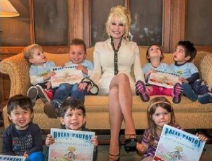 DollyParton sits on a couch with several small children all holding up a book about Dolly Parton.