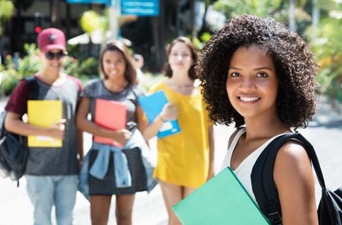 A girl stands with a backpack and binder smiling brightly at the camera. Her three friends stand in the background.