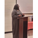 A woman wearing a pearl necklace is speaking at a podium. 