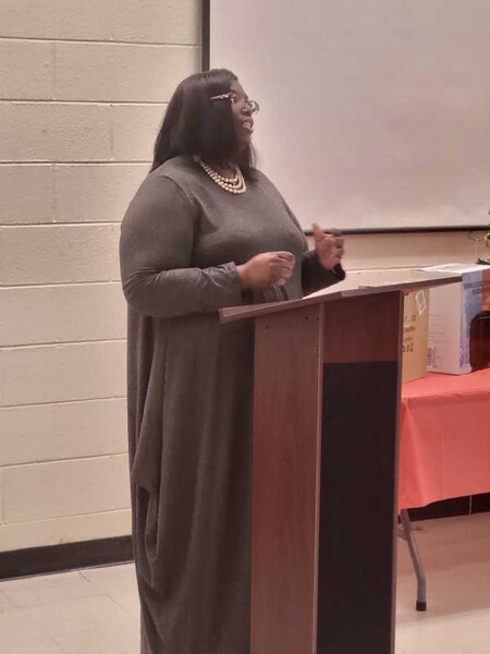 A woman wearing a pearl necklace is speaking at a podium. 