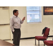 A gentleman is talking with his hands at the front of a room. 