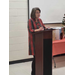 A woman who in a plaid shirt addresses the room from the podium. 