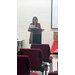 The woman in plaid stands at the podium and reads from a piece of paper. 