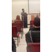 A woman in black-rimmed glasses addresses the room of people from the podium. 