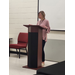 A woman wearing a lanyard talks behind a podium.