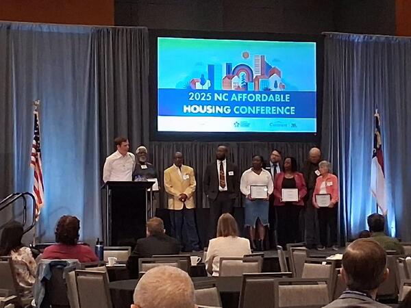 Nine people stand on the stage in front of a screen that says 2025 NC Affordable Housing Conference.