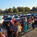  People in line to get bags of food, in front of a parking lot full of cars. 
